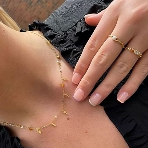 Nærbilde av en kvinnes hals og hånd. Hun bærer et tynt gullkjede med små hengende dråper, og flere enkle gullringer på fingrene.