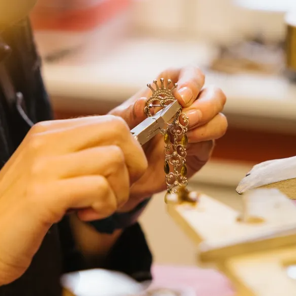 Nærbilde av hender som bruker verktøy (tenger) til å forme eller montere et intrikat metallkjede ved en gullsmedbenk.