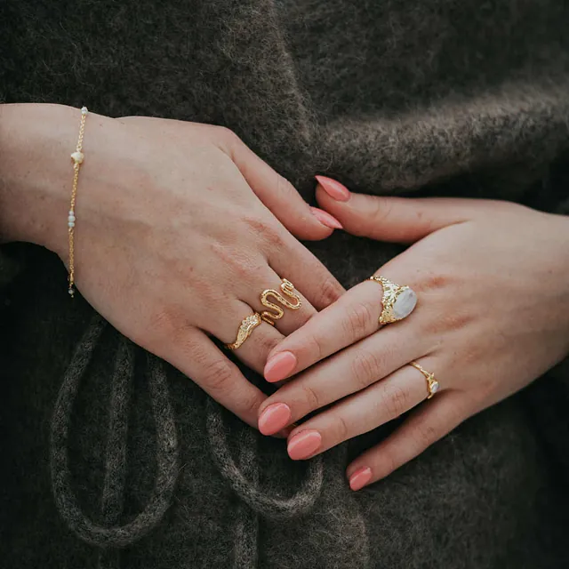 Nærbilde av hender med lys neglelakk som hviler på en mørk strikkejakke. Hendene er pyntet med flere gullringer, inkludert en med en stor perle, og et tynt armbånd.