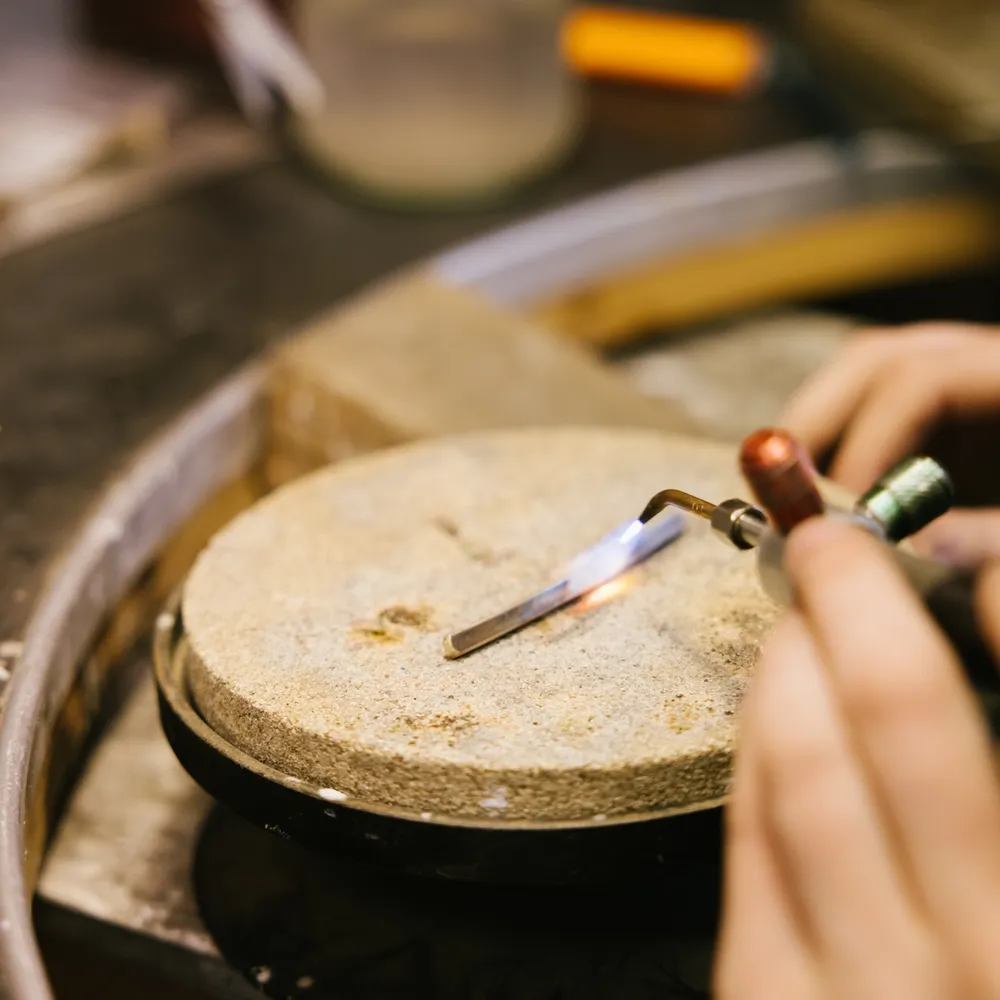 Hender som bruker en gassbrenner til å varme opp en metalltråd som holdes med pinsett på en rund loddeplate.