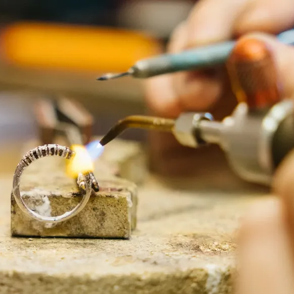 Nærbilde av en gullsmed i arbeid. En hånd bruker en gassbrenner med åpen flamme til å varme opp et smykke som holdes fast med en tang på en arbeidsbenk.