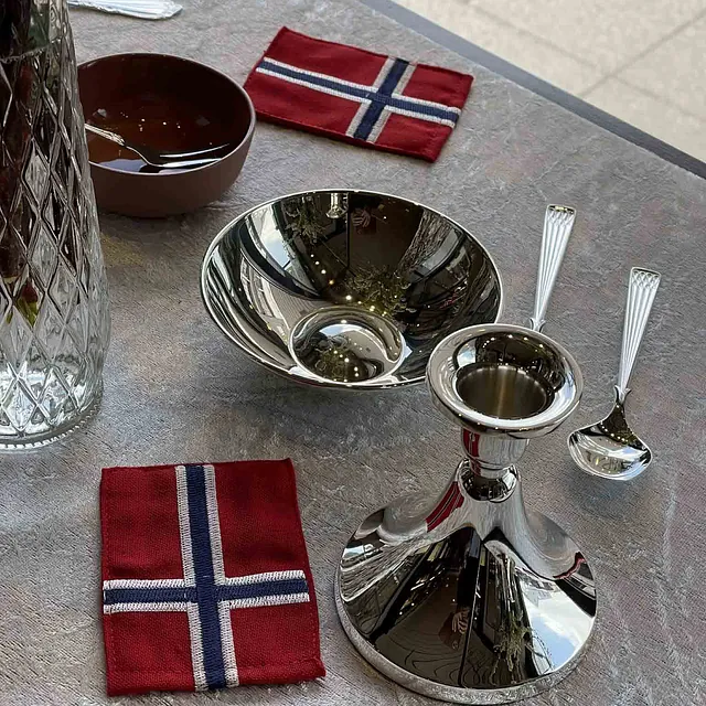 Borddekking med lysestake og en skål i blankt sølv, en sølvskje, og servietter med det norske flagget. I bakgrunnen står en krystallkaraffel.