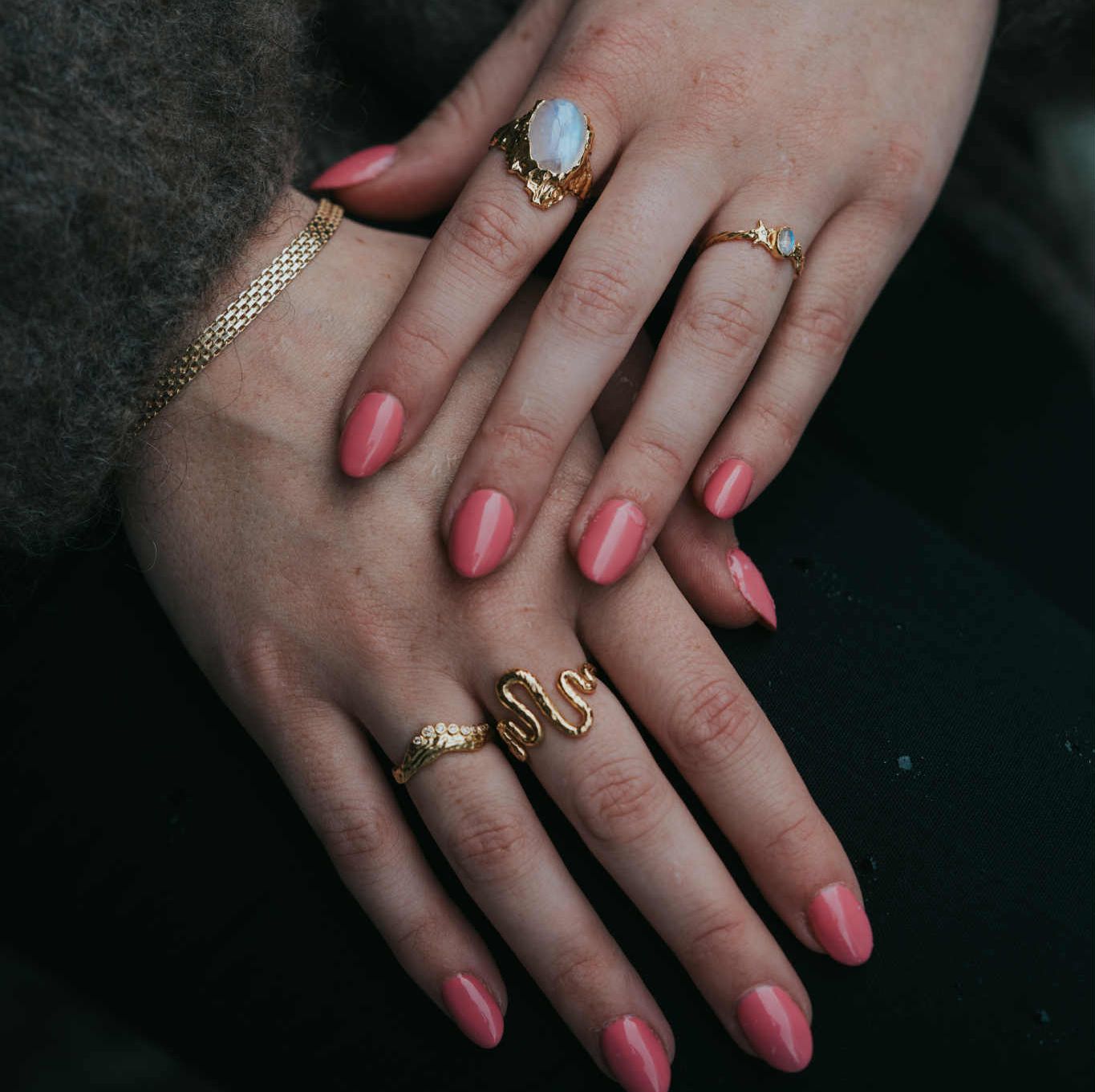 Hender pyntet med flere gullringer, inkludert en som former en slange og en med en stor, lys stein, samt et nettingarmbånd i gull.